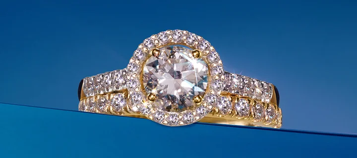 Złoty pierścionek z dużym okrągłym diamentem otoczonym mniejszymi brylantami na niebieskim tle