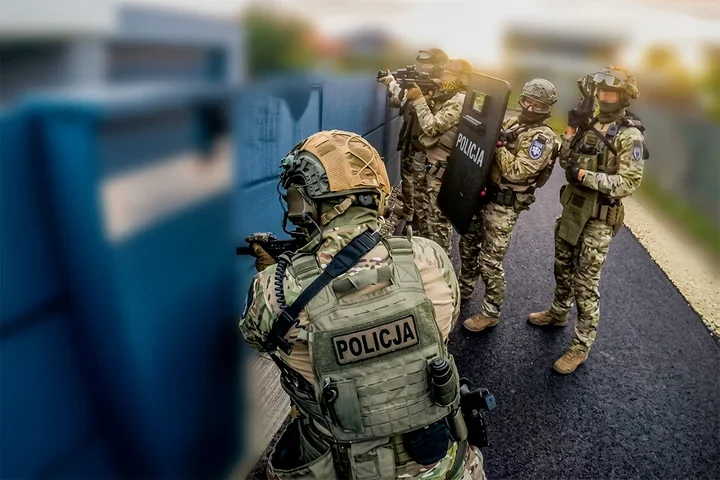Grupa policjantów w pełnym rynsztunku taktycznym zabezpiecza teren przy niebieskim ogrodzeniu.