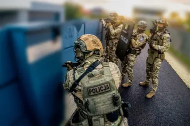 Grupa policjantów w pełnym rynsztunku taktycznym zabezpiecza teren przy niebieskim ogrodzeniu.