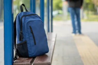 Niebieski plecak pozostawiony na ławce na przystanku autobusowym, w tle niewyraźna postać stojącej osoby.